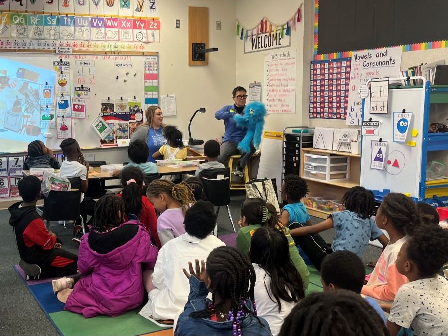 Two WoodmenLife volunteers, both wearing blue shirts, sit in front of a group of students. One of the volunteers holds a fuzzy blue puppet up for all the children to see.