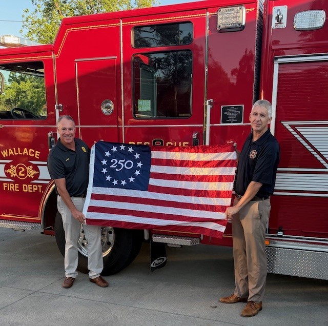 Two men hold a commemorative Betsy Ross 250 U.S. flag in front of a fire truck.