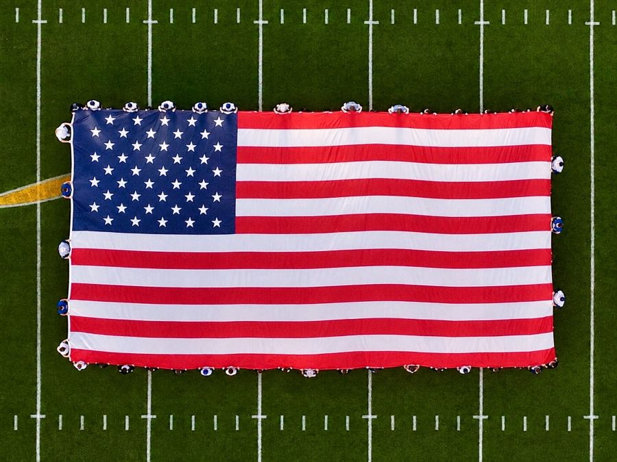 Photo is a drone image looking down on an oversized American flag displayed on a football field.