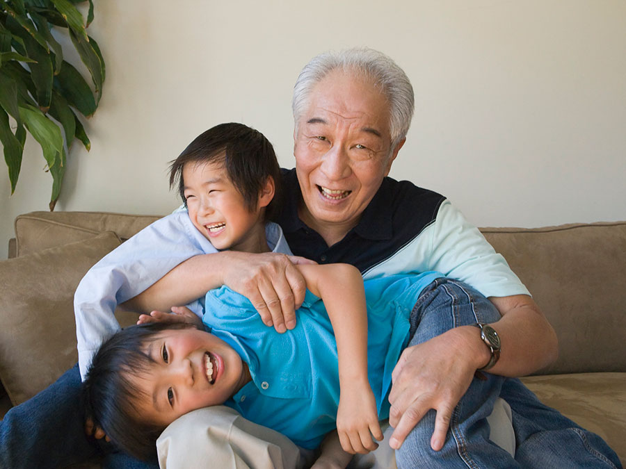 An older man sits with two younger boys — they appear to be a grandfather and two grandsons. One boy is sitting beside the man, leaning into him, and the other boy is laying across the man's lap. Their arms are around each other, and everyone is smiling.