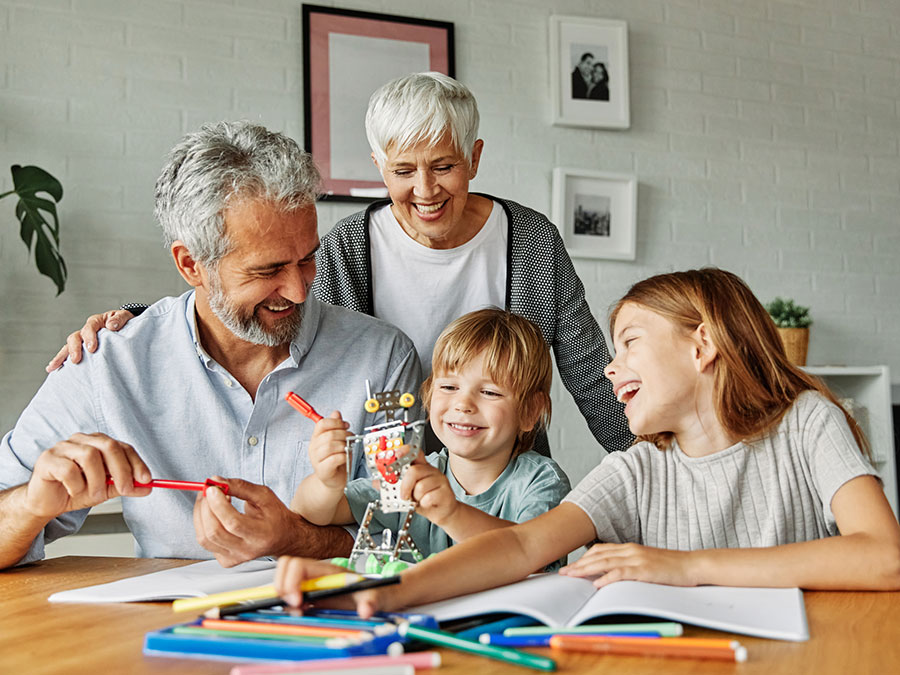 An older man and woman sit with a younger girl and boy at a table — they appear to be grandparents and grandchildren. On the table in front of them are books, colored pencils, and toys. Everyone is smiling.