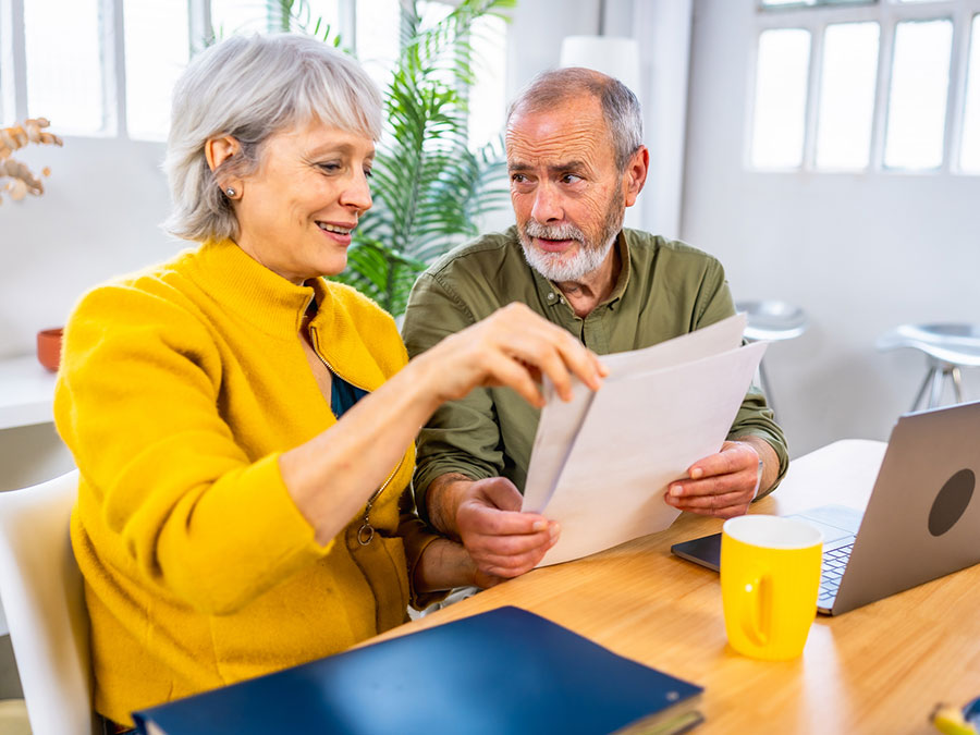 Stock photo shows an older couple sitting at a table. There is an open laptop and a coffee cup on the table in front of them. They are holding paperwork that they're looking over.