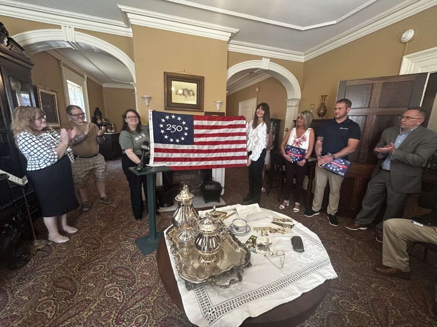 Two women hold open a commemorative Betsy Ross 250 U.S. flag as a group of people look on and clap.