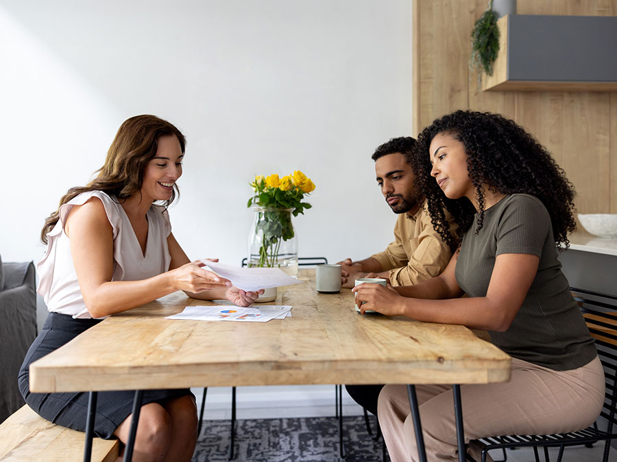 Stock photo shows a woman sitting on one side of a table, and a man and woman sitting on the other. The woman is holding a piece of paper and explaining it to the couple. There is also a vase with flowers on the table.