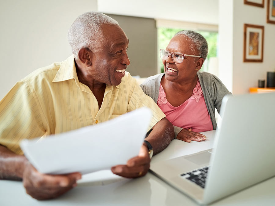 An older couple sit at a desk. There is a laptop on the table in front of them. The man is holding a piece of paper. They are both smiling and looking at each other.