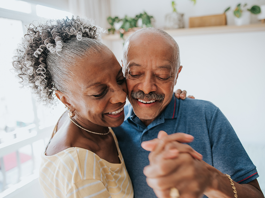 Stock photo shows a close-up view of an older man and woman dancing. They are standing close, hands joined and cheeks touching. They are both smiling.