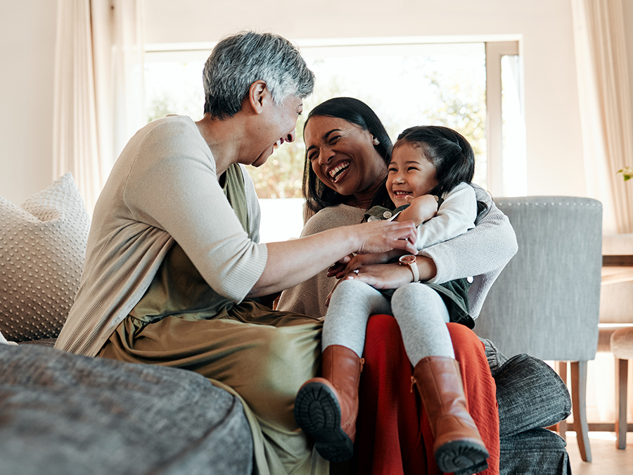 Stock photo shows three people sitting together on a couch. An older woman sits beside a middle-age woman, and a young girl sits on the middle-age woman's lap. They are all smiling and embracing. Behind them is a table with chairs and a window.