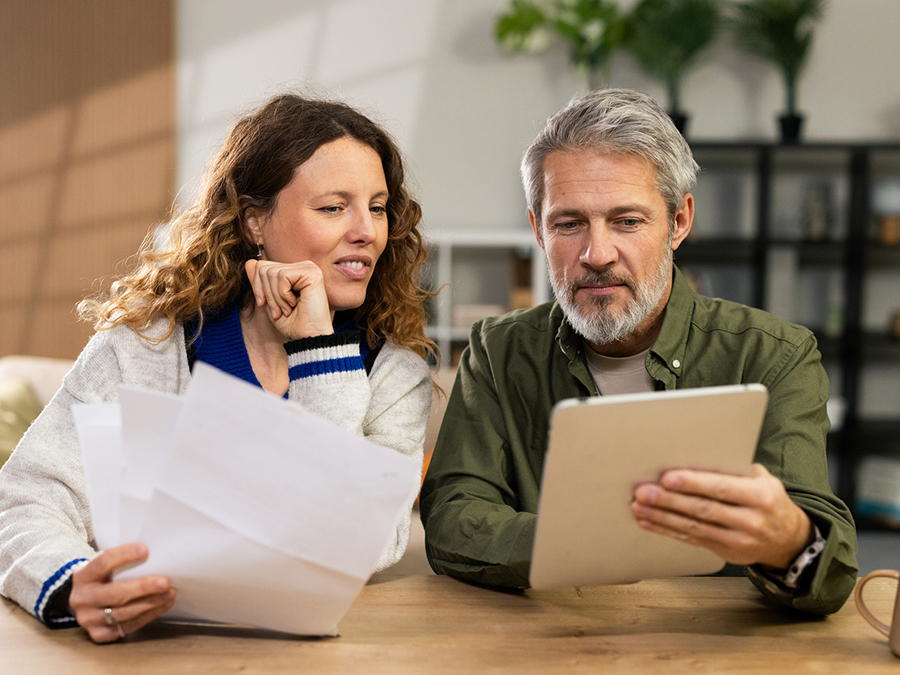 Stock photo shows a man and a woman sitting together at a table. The man is holding a tablet, which they are both looking at. The woman is holding a piece of paper.