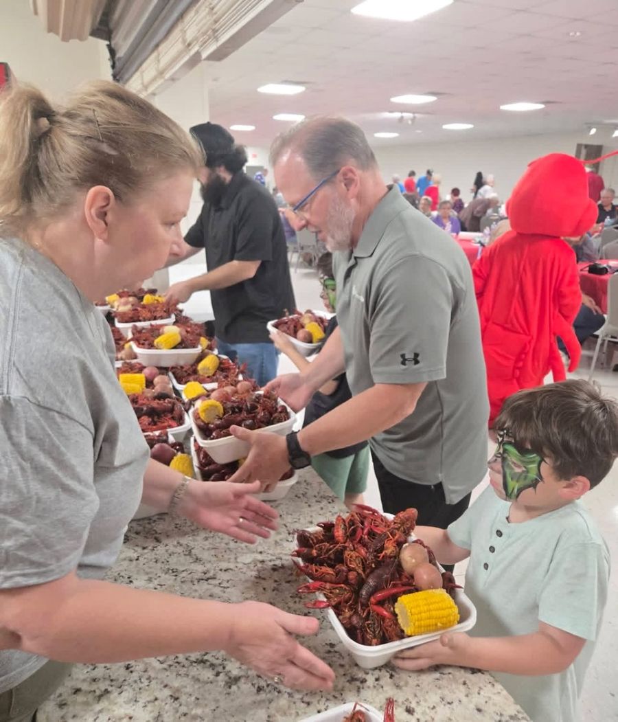 Photo shows a woman handing a plate to a young boy. The plate is full of crawfish, potatoes, and corn-on-the-cob. The boy's face is painted. They are standing on either side of a long counter filled with plates, ready to be distributed.