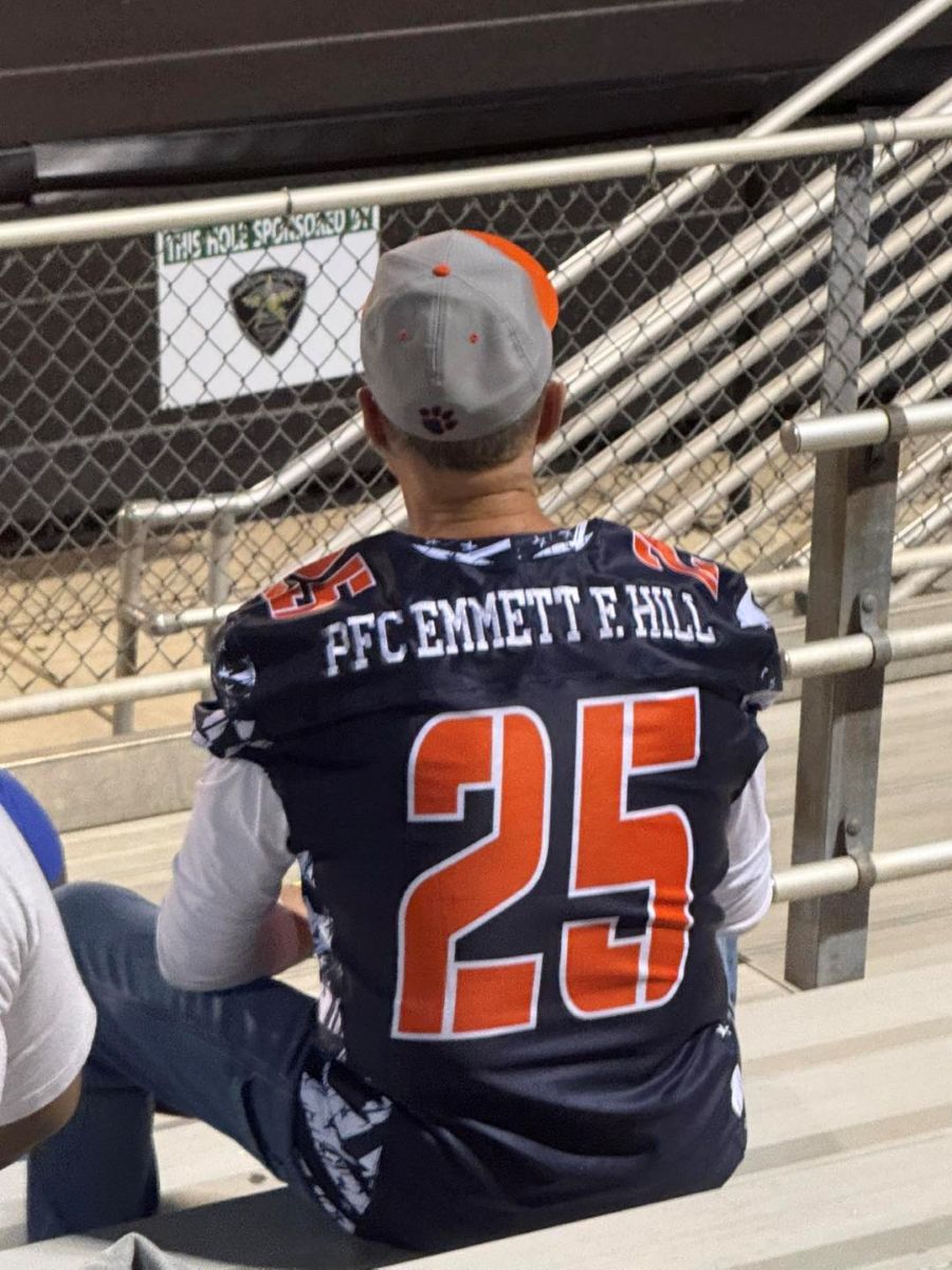 Photo shows the back of someone who is seated on a football stadium bleacher, watching the game. The person is wearing a replica jersey that is navy blue with orange and white lettering. This person's jersey is No. 25 and has the name PFC Emmett F. HIll.