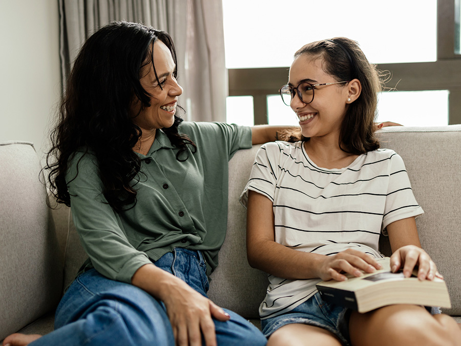 Stock photo shows an adult woman and a teen girl sitting together on a couch. The woman has her arm around the girl's shoulders, and both are smiling at each other. There is a window behind them.
