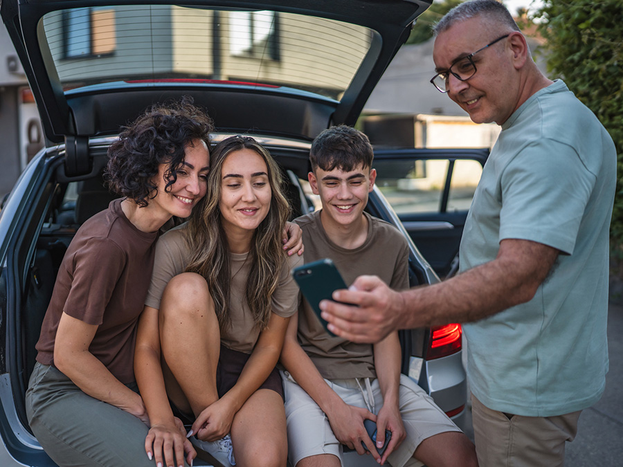 Stock photo shows a family of four -- an adult man and woman and a teen boy and girl -- sitting in the back of a hatchback car. The man is holding a cellphone out for the other three to look at.