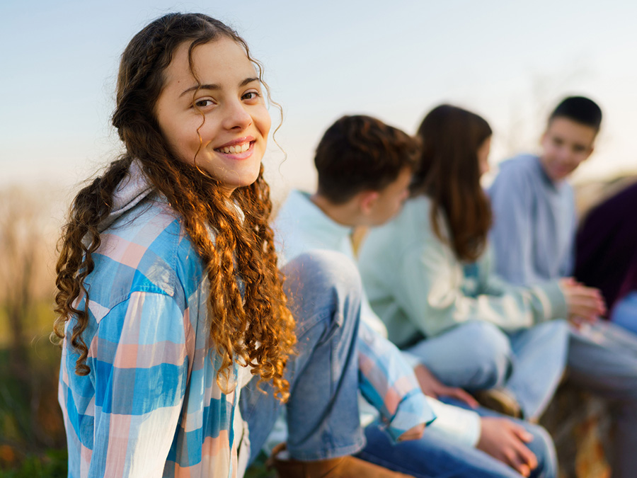 Stock photo shows a teen girl sitting outside, smiling for the camera. Behind her are four other people talking among themselves. There is blue sky behind them.