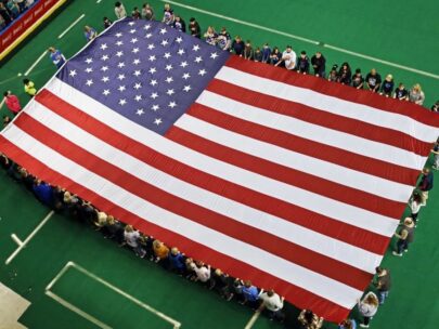 A 30-foot-by-60-foot American flag is seen from above. The flag is being presented on the green turf of an indoor football field. More than 70 volunteers are standing around the flag and holding it.