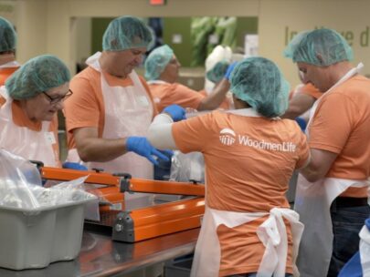 A group of WoodmenLife associates — all wearing orange shirts, hair nets and plastic aprons — volunteer together at the Food Bank for the Heartland during Strike Out Hunger month.