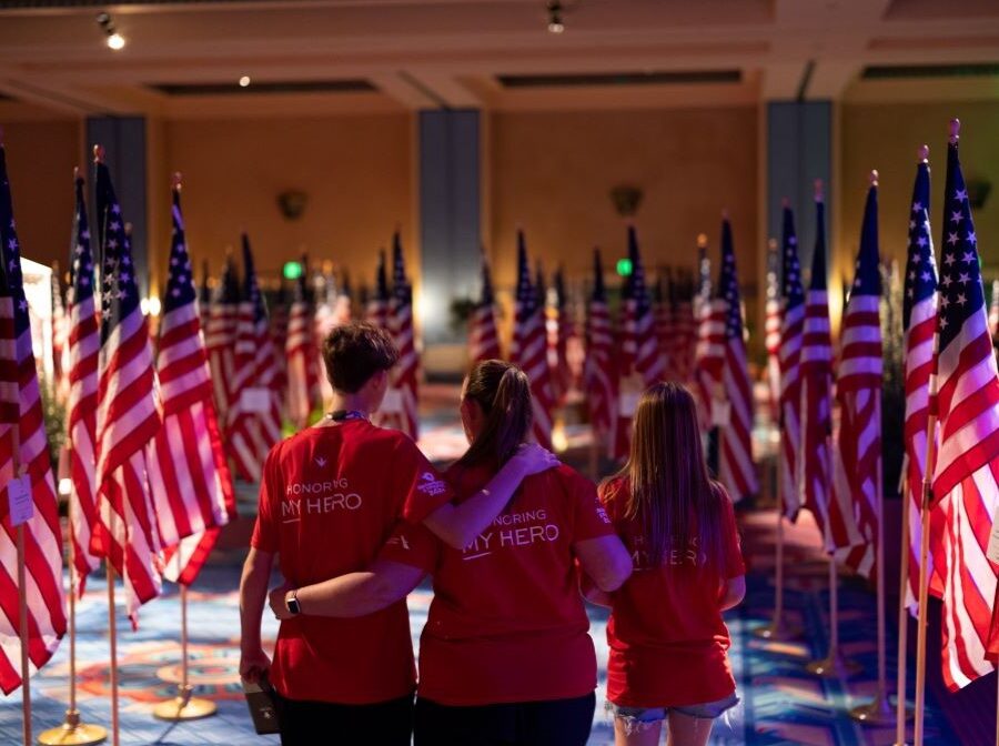 Photo shows the backs of three people, their arms around each other. They are standing in a dim room filled with American flags. They appear to be soaking in the significance and emotion of the moment.