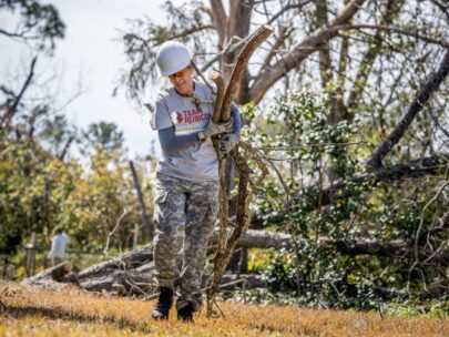 Photo shows a volunteer wearing a gray Team Rubicon T-shirt, camo pants, and a white hard hat. She is carrying large tree branches. Behind her are other trees, both upright and on the ground.