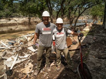 Photo shows two men. They are both wearing grey T-shirts that say "Team Rubicon Disaster Response" and their names. They are wearing white hardhats and work gloves, and one man is holding a shovel. They are standing outside along a waterway. Behind them is wreckage from a recent natural disaster.