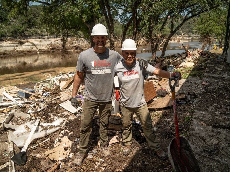 Photo shows two men. They are both wearing grey T-shirts that say "Team Rubicon Disaster Response" and their names. They are wearing white hardhats and work gloves, and one man is holding a shovel. They are standing outside along a waterway. Behind them is wreckage from a recent natural disaster.