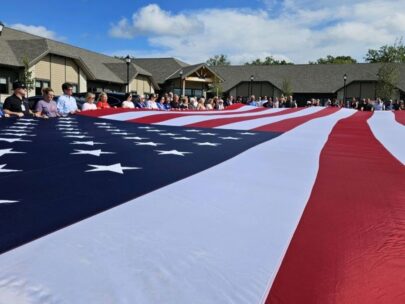 Photo is looking across an oversized American flag. People are standing around the edges of the flag, holding it up. There is a building and blue sky in the background.