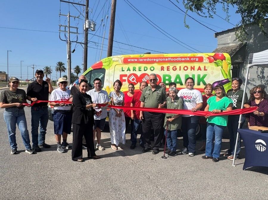 Photo shows 18 people holding a red ribbon in front of a colorful cargo van that says "Laredo Regional Food Bank" on the side. A woman in the middle is holding oversized scissors to cut the ribbon.