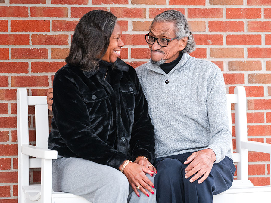 WoodmenLife member Bessie Pierce and her husband, William, are seated together on a bench. She is wearing a black coat, and he is wearing a gray sweater. They are looking at each other and smiling.