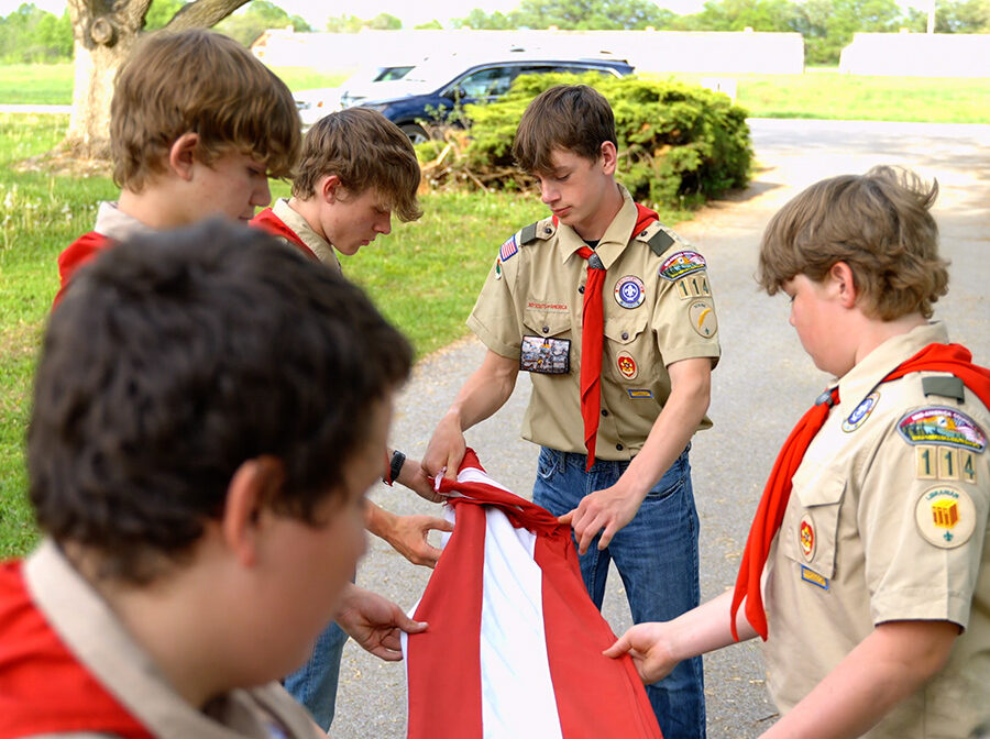 Six Scouts hold the edges of an American flag, working together to fold it.