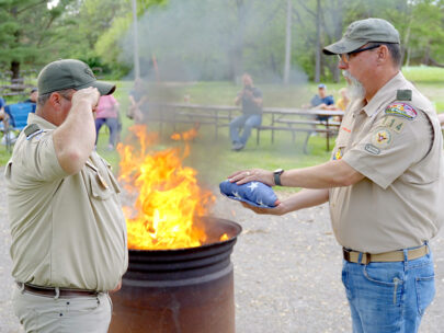 One Scout leader holds out a folded U.S. flag, while a second Scout leader salutes. Behind them is a fire in a fire pit.
