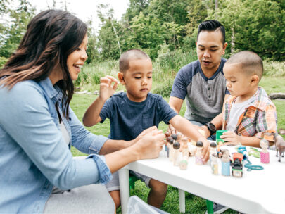 Stock photo of a mom, dad and two young boys sitting at a table outside. They are playing with small figurines on the table.