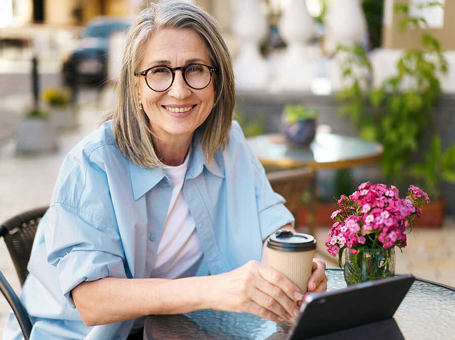 Image is a stock photo of a woman sitting at a table. She is holding a coffee cup. On the table in front of her is a tablet propped upright. There is also a small vase with flowers on the table.