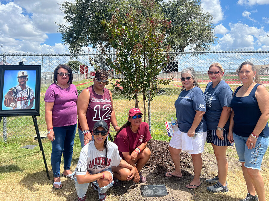 Seven women pose for a picture around a newly planted tree. Next to them on a stand is a framed picture of the young WoodmenLife member who passed away. Behind the group is a fence, on the other side of which is an athletic field.