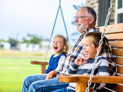 This image is a stock photo of an older man sitting on a porch swing with a young child — likely his grandchildren — on either side of him.