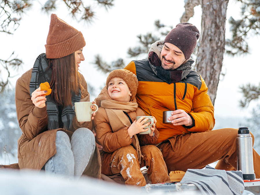 Image is a stock photo of a family sitting outside in cold weather. Family consists of a mom, a dad and a child sitting between them. The family is dressed warmly, wearing coats, hats and scarfs. They are also enjoying a warm beverage and cookies.