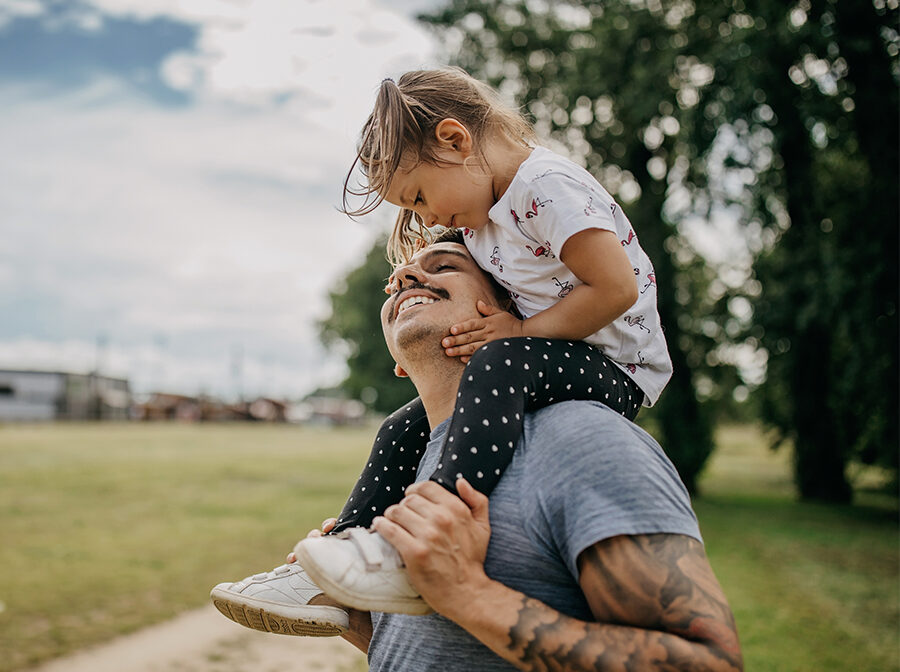 Image is a stock photo of a man holding his toddler daughter on his shoulders. He is looking up at her, and she is looking down at him and holding his face. They are outside, and there is grass, trees, and sky behind them.