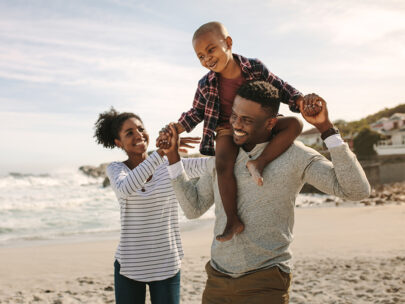 Image is a stock photo of a family walking on a beach. The dad holds their young son on his shoulders, while the mom walks alongside, helping the boy stay up.