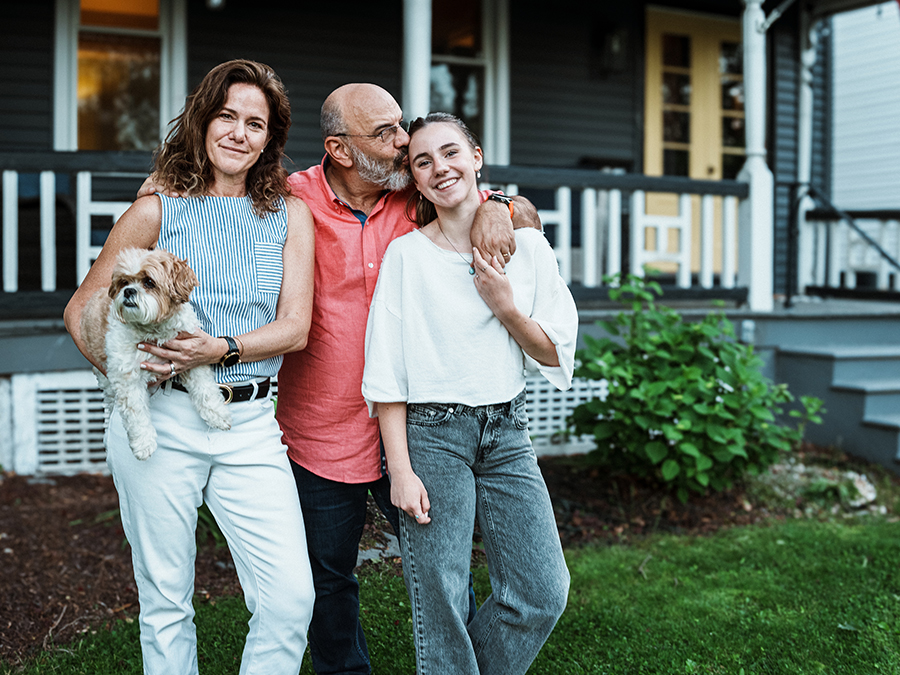 Stock photo shows a family of three: a woman, a man, and a teenage girl. The woman is holding a small white dog. They are standing outside, with the front door and front porch of a house behind them.