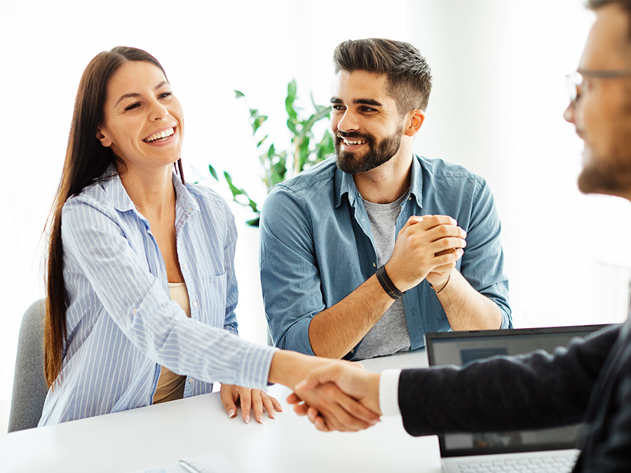 Stock photo shows a man and a woman sitting at a table across from a man. The woman is shaking hands with the man across the table. All three people are smiling.