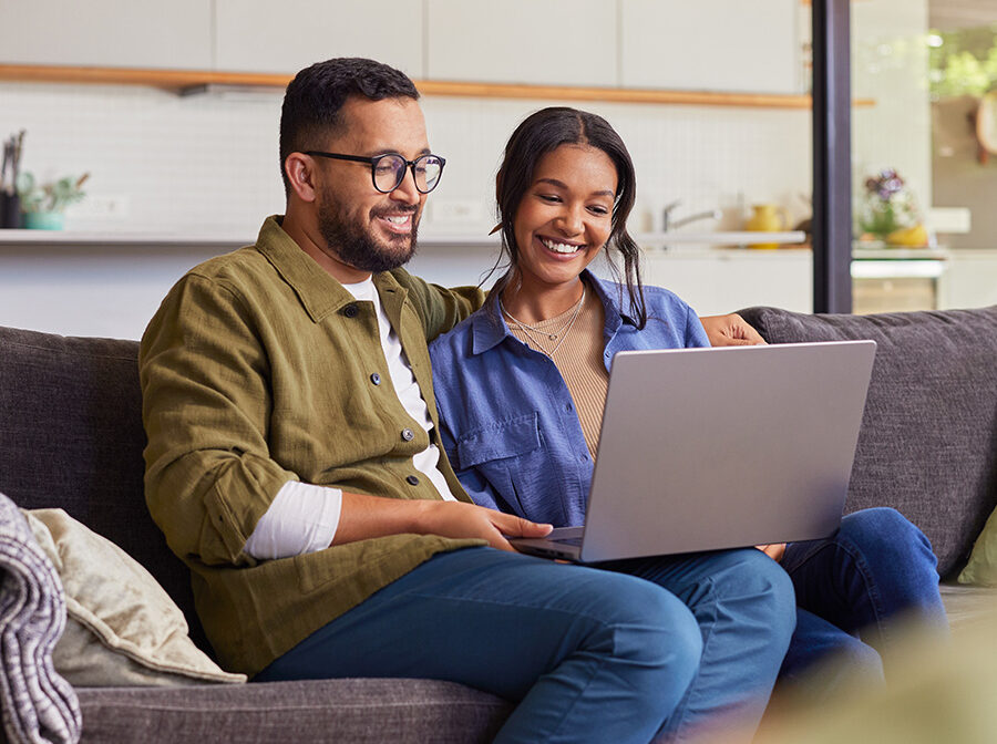 Stock photo shows a man and a woman sitting on a couch looking at a laptop screen. They are both smiling.