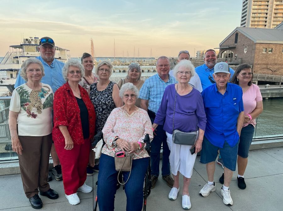 A group of 13 people stand together for the photo. They are outdoors, and behind them is water with boats.