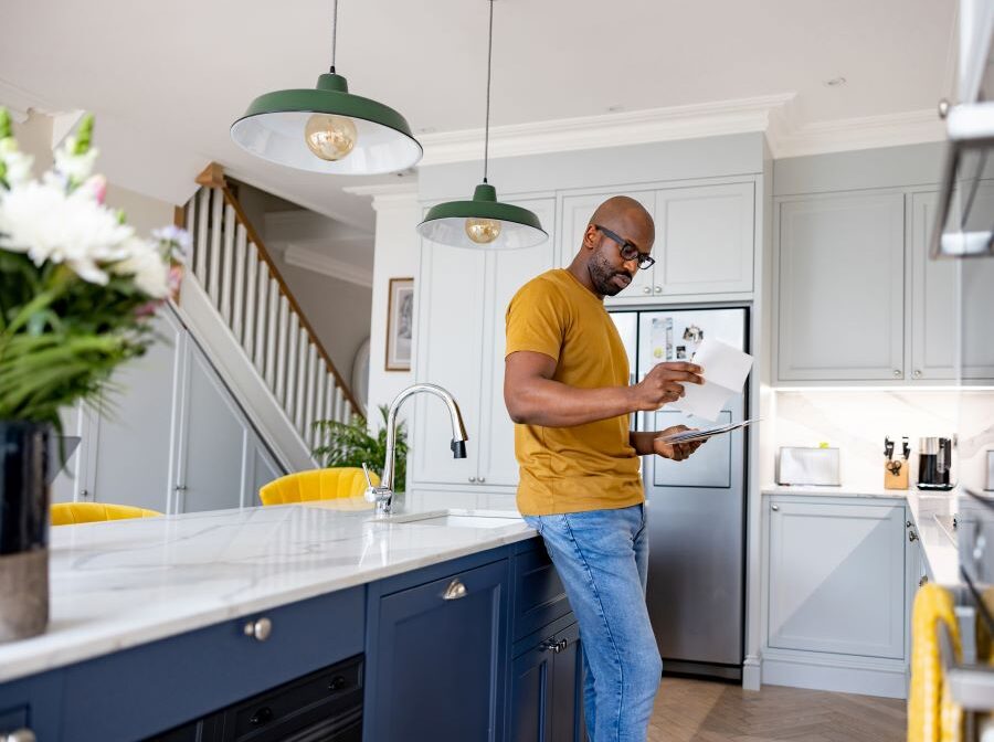 Stock photo shows a man standing in a kitchen, leaning against a counter and looking over a piece of paper.