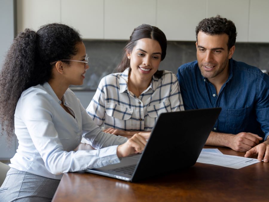 Stock photo shows a man and woman sitting at a table with a life insurance professional, who is pointing at a laptop screen. The man and woman are looking on, smiling, and listening to the professional explain.