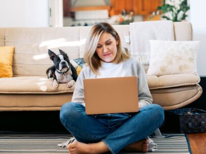 Stock image shows a woman sitting on the floor with a laptop on her lap. She is sitting against a couch, and there is a small dog laying on the couch, looking over her shoulder at the laptop.