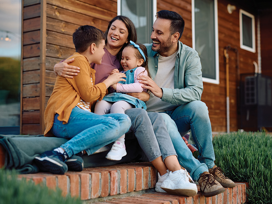 Stock photo shows a family — a mom, a dad, a young boy, and a young girl — sitting on a home's front steps. The daughter is sitting on the mom's lap, and everyone has their arms around each other, smiling and laughing.