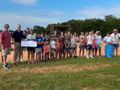 A group of 28 people — a mix of adults and children — stand together for the photo. People on the left side of the group are holding a WoodmenLife-branded oversized check. It is made out to Cargo Ranch in the amount of $4,000. The group is standing outside — in front of them is green grass, behind them is an area of dirt or sand, and above them is a blue sky.