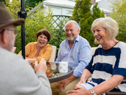 Four people sit outside around a table. They are smiling and laughing. On the table is a coffee cup, and behind the people are green shrubs and a fence.