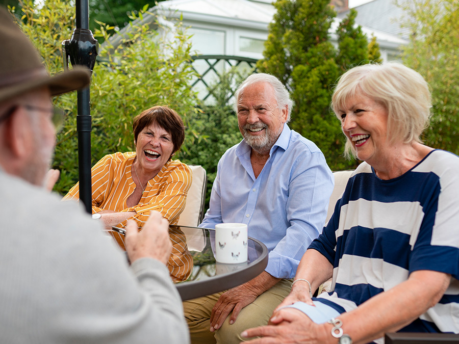 Four people sit outside around a table. They are smiling and laughing. On the table is a coffee cup, and behind the people are green shrubs and a fence.