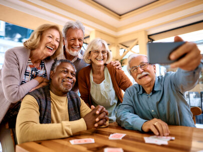 Stock photo shows a group of five older adults. They are standing together posing for a picture, as one of the men holds out a cellphone to take a selfie. On the table in front of them are playing cards.