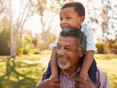 Stock photo shows a young boy sitting on the shoulders of an older man. The boy's arms are around the man's neck, and both are smiling. They are outside, and you can see behind them sunlight, trees, and grass.