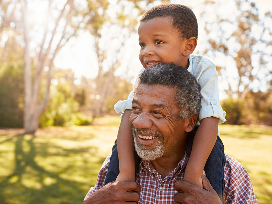 Stock photo shows a young boy sitting on the shoulders of an older man. The boy's arms are around the man's neck, and both are smiling. They are outside, and you can see behind them sunlight, trees, and grass.