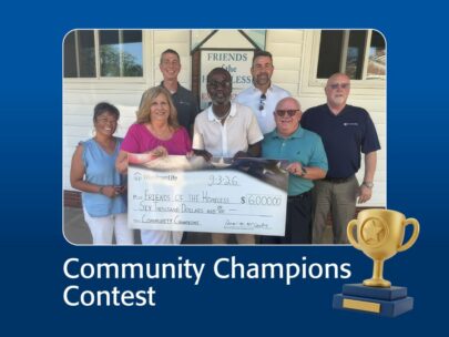 Graphic includes a dark blue background and the words "Community Champions Contest" across the bottom. In the middle of the graphic is a photo. The photo shows seven people standing together holding an oversized WoodmenLife-branded check. The check is made out to Friends of the Homeless in the amount of $6,000. In the lower right corner of the graphic is an illustration of a gold trophy.